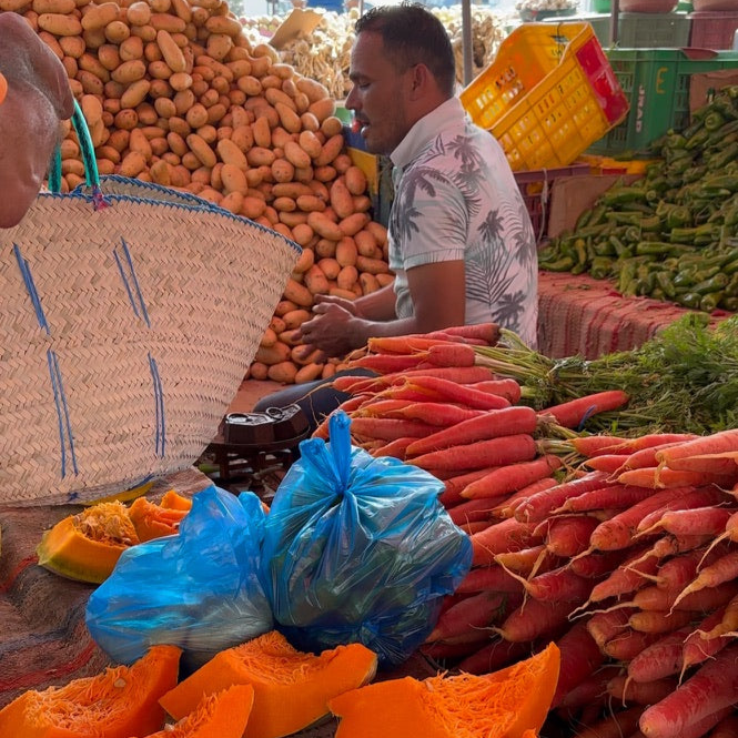 A market vendor’s table full of fresh carrots, potatoes, and squash next to Vivian’s woven basket waiting to be filled — everyday scenes that shape artisan goods.
