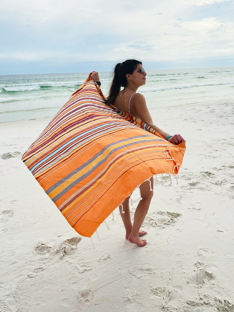 Woman on the beach wrapped in vibrant orange oversized fouta bath towel waving in the wind.
