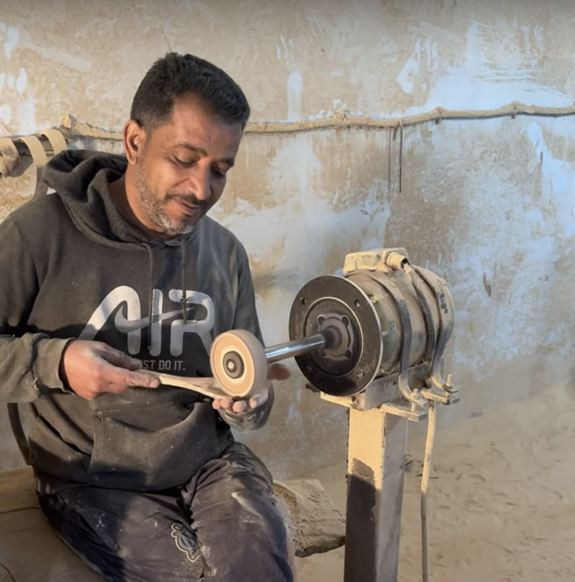Moncef seated at a sander inspecting and smoothing an olive wood spoon cupped in his hand — final touches on a timeless handcrafted gift.